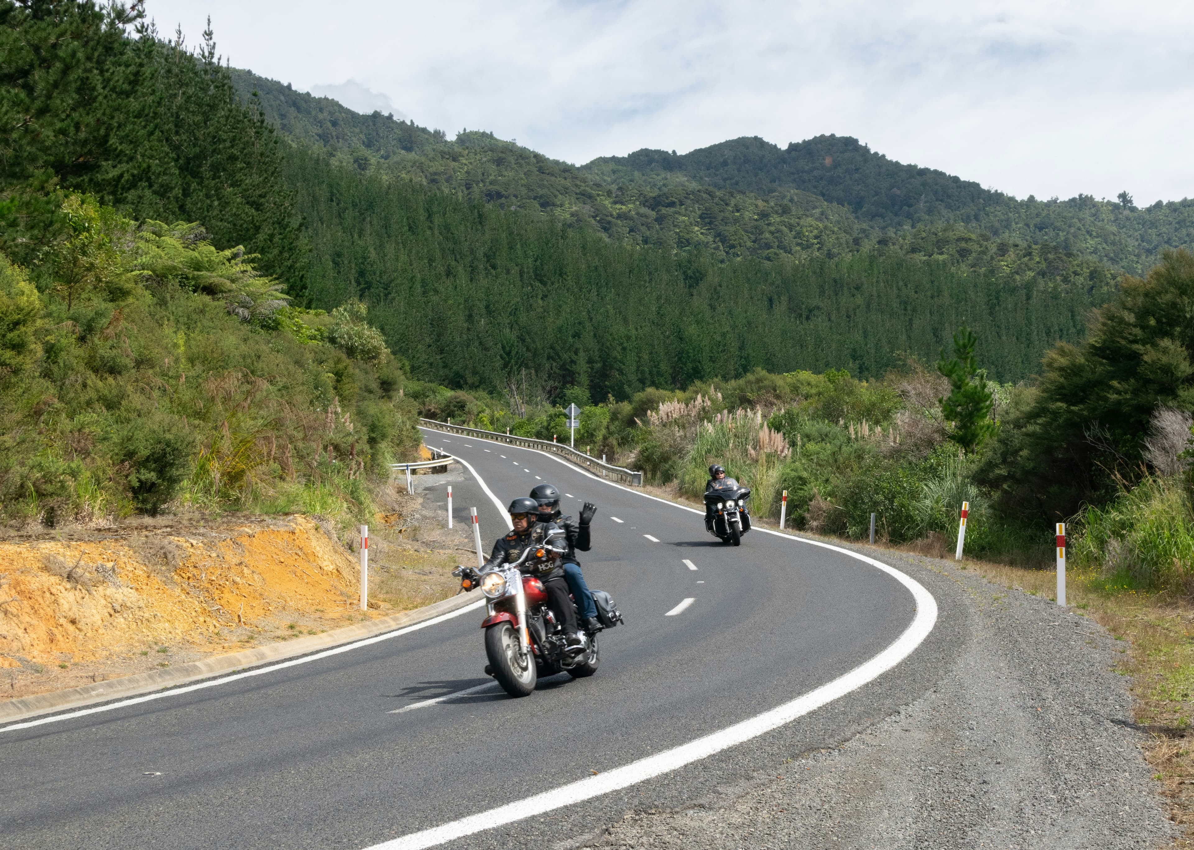 Riders waving as they come through a bend on a coastal road lined with green hills